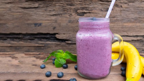 Purple aronia smoothie in glass jug with bananas and blueberries.