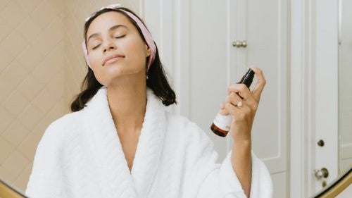 Woman in bathrobe spraying her face with a beauty product